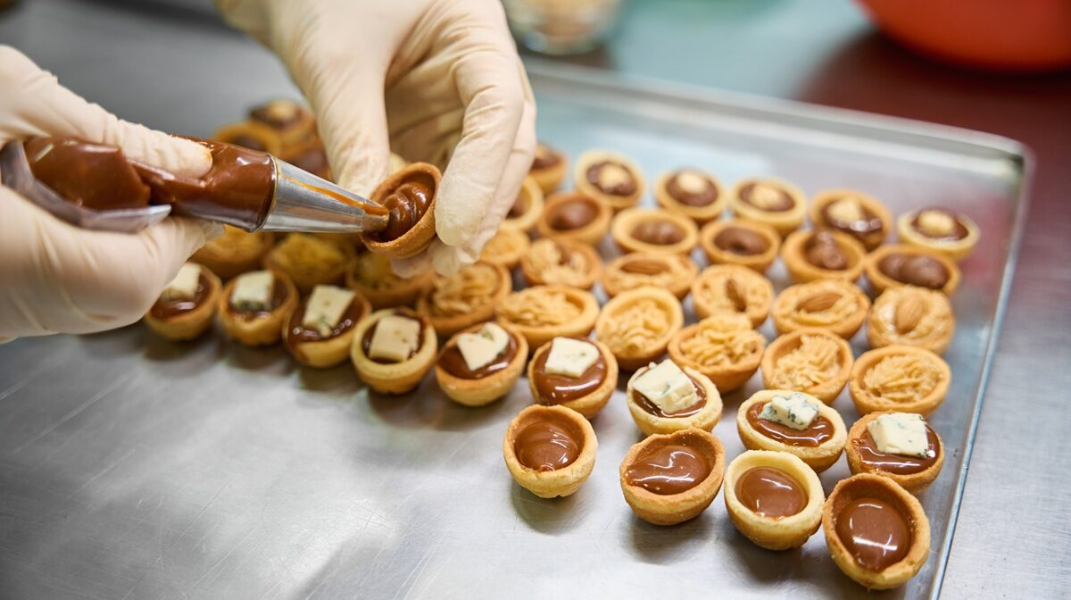 Close-up baker hands holding pastry bag with chocolate fudge and filling the sweet pastry nuts, cookie halves decorated with cheese and nuts lying on the tray