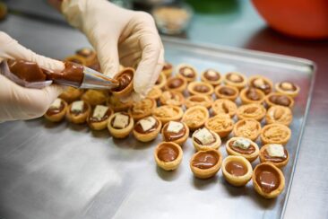 Close-up baker hands holding pastry bag with chocolate fudge and filling the sweet pastry nuts, cookie halves decorated with cheese and nuts lying on the tray