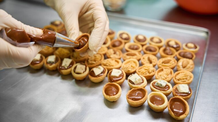 Close-up baker hands holding pastry bag with chocolate fudge and filling the sweet pastry nuts, cookie halves decorated with cheese and nuts lying on the tray