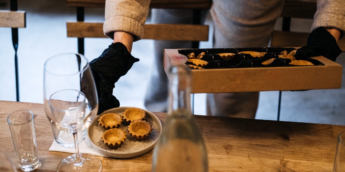 A caterer in black gloves meticulously places fresh gourmet tartlets onto a plate at a wine tasting event, highlighting attention to detail.