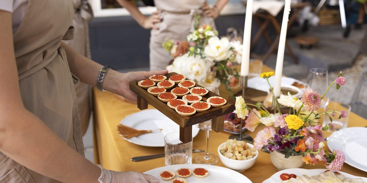 Wedding. Banquet. Chairs and a table for guests, decorated with candles, are served with cutlery and crockery and covered with yellow tablecloth. Waiter setting table stands on a green lawn in the backyard banquet area